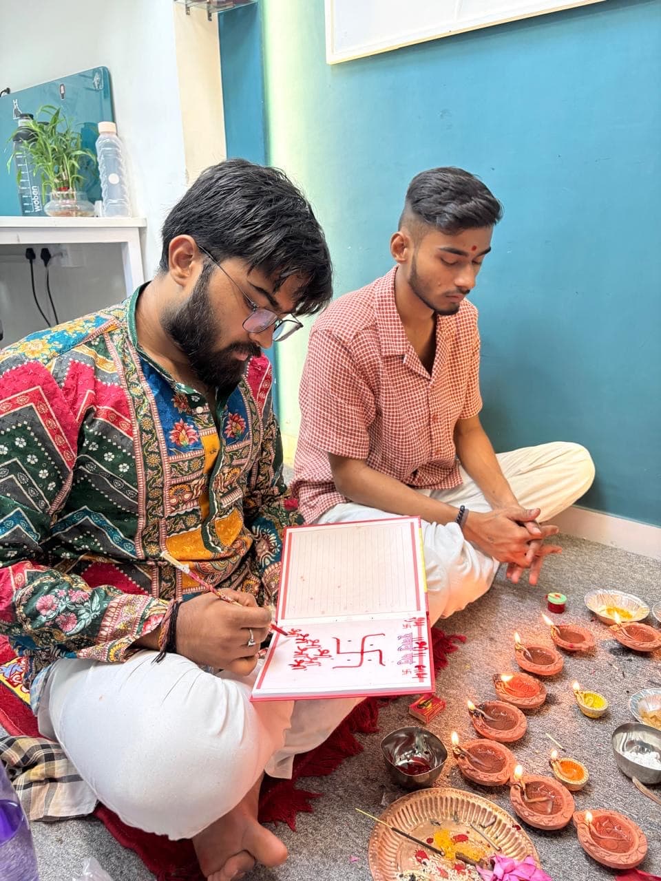 Two members of the Stucare team performing the Diwali chopda pujan, writing a Swastik and Shubh-Labh in a ceremonial ledger beside rows of lit diyas.